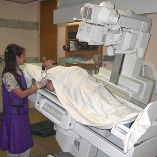 Medical professional operates an X-ray machine for a patient lying on a table, covered by a white sheet.