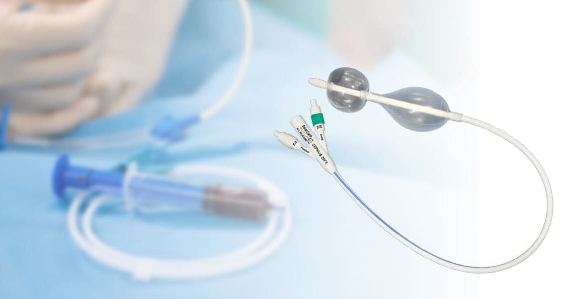 A Foley catheter with dual balloons and multiple ports is shown; medical tubing and a syringe are visible in the blurred background.