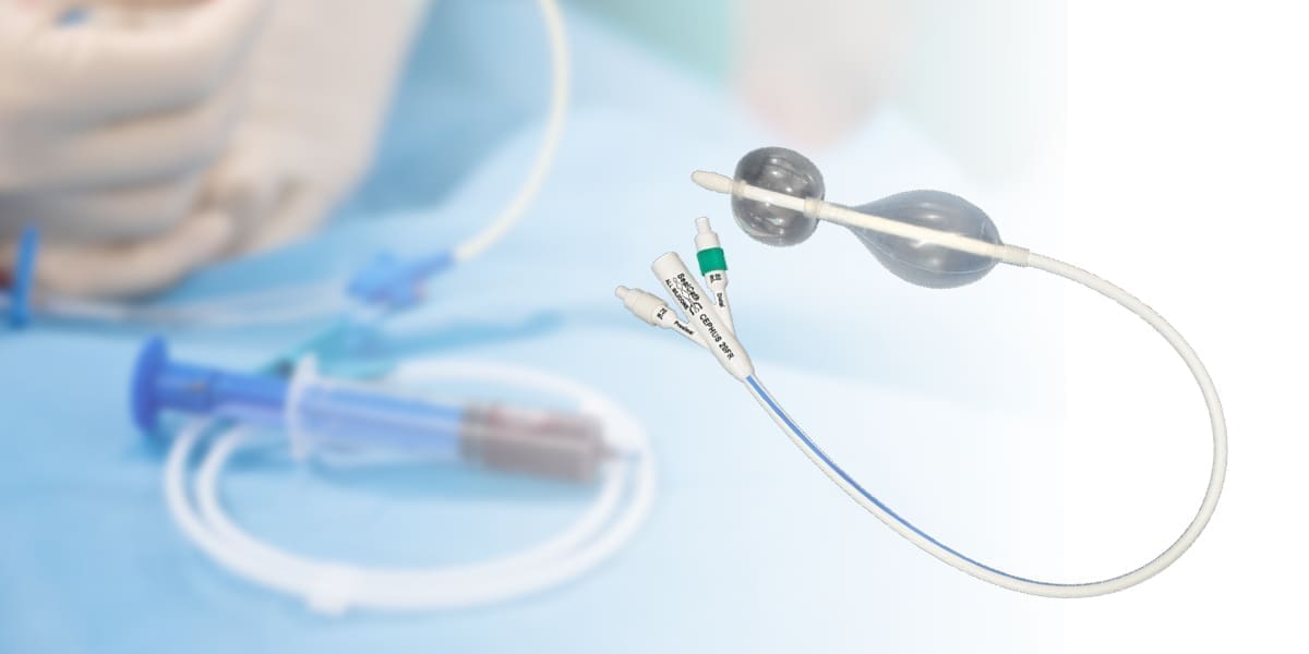 A Foley catheter with dual balloons and multiple ports is shown; medical tubing and a syringe are visible in the blurred background.