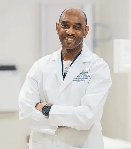 A man in a white lab coat stands with arms crossed, smiling at the camera in a clinical or hospital setting.